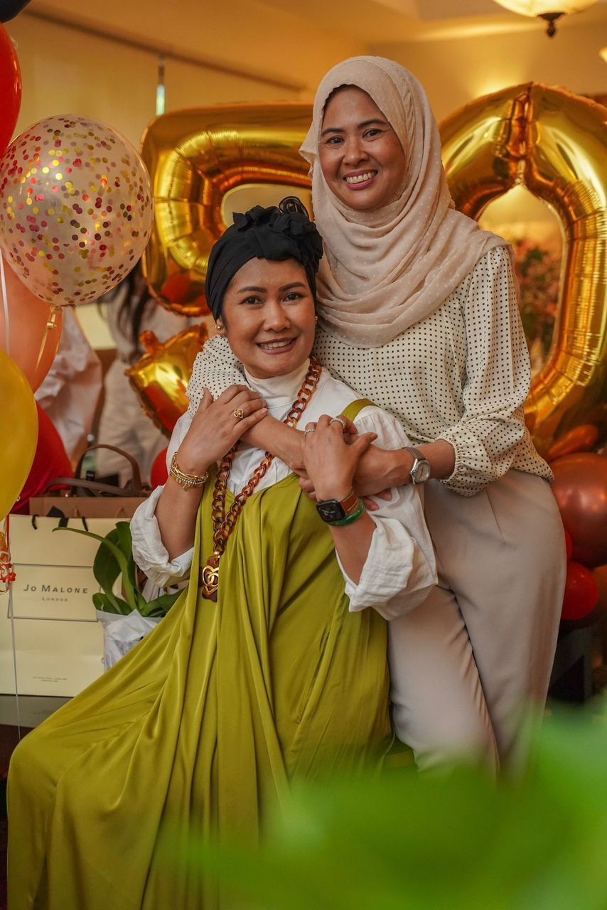 Two women are standing and sitting close together, surrounded by festive balloons and gift bags.