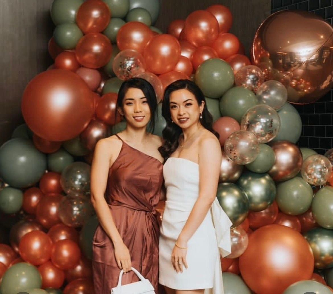 Two women in elegant dresses posing in front of a colorful balloon backdrop.