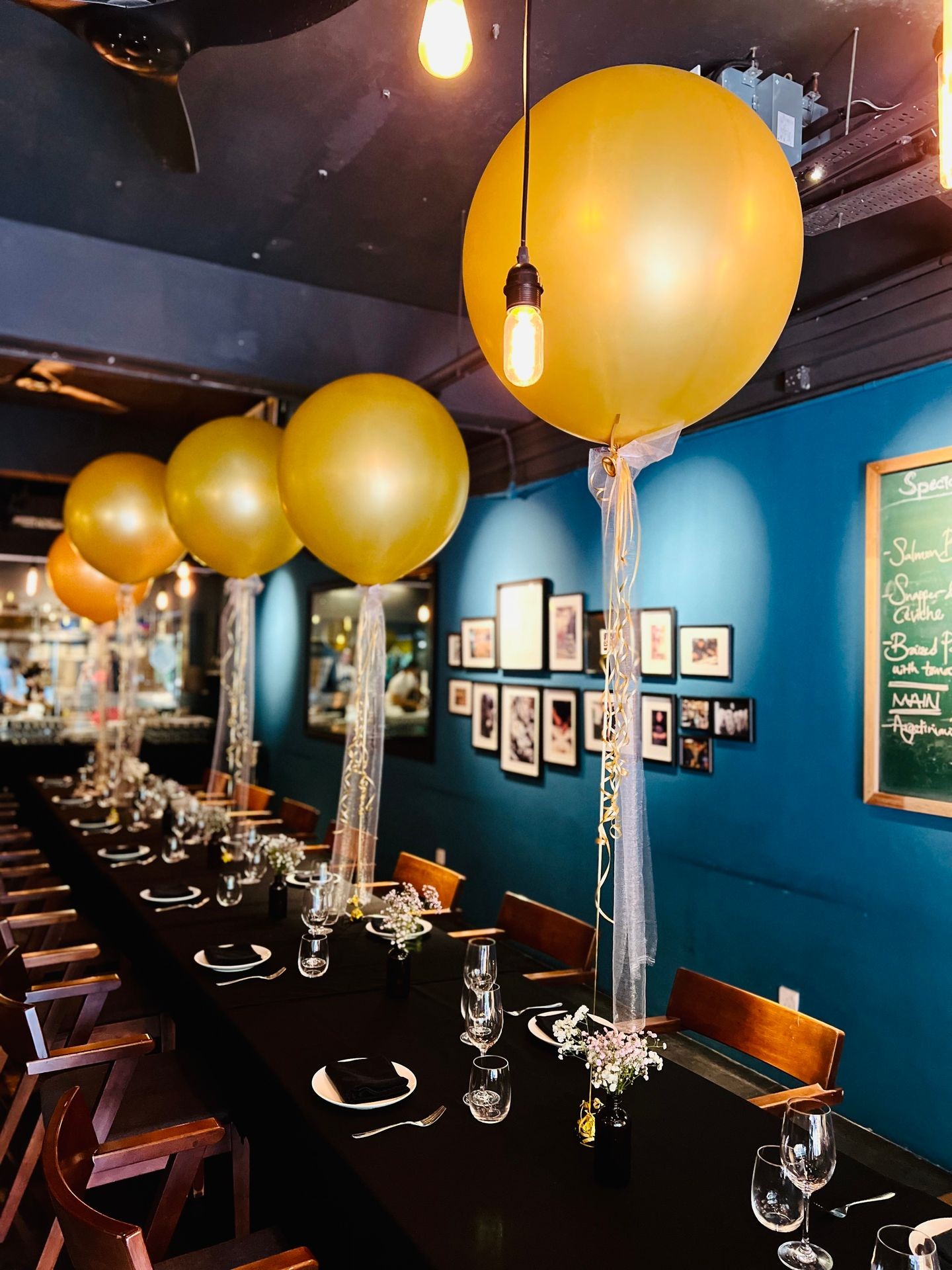 Restaurant table setup with large gold balloons and elegant place settings, blue wall in background.